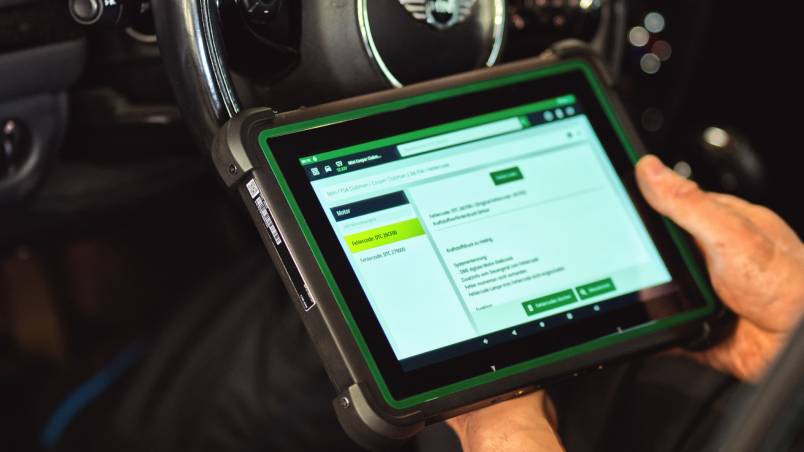 Das Bild zeigt eine Person im Auto, die mit einem Diagnosegerät den Fehlerspeicher ausliest. Auf dem Tablet sind Informationen zu möglichen Fehlern sichtbar.