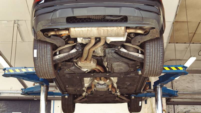 Das Bild zeigt ein Auto, das auf einer Hebebühne in einer Werkstatt angehoben ist. Der Unterboden mit Auspuffanlage und Fahrwerk ist gut sichtbar.