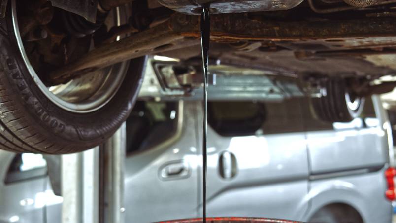 Gezeigt wird ein Ölwechsel. Es läuft dunkles Motoröl aus der Ölwanne des Autos in einen roten Auffangbehälter. Eine Hand hält den Behälter, während das Fahrzeug von unten sichtbar ist.