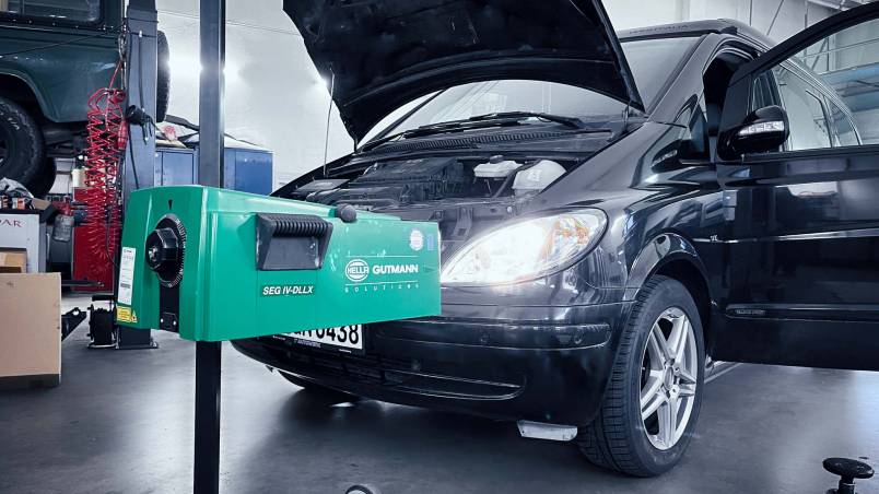 Das Bild zeigt ein Auto in einer Werkstatt mit eingeschaltetem Scheinwerfer. Vor dem Fahrzeug steht ein grünes Prüfgerät zur Kontrolle und Einstellung der Fahrzeugbeleuchtung.