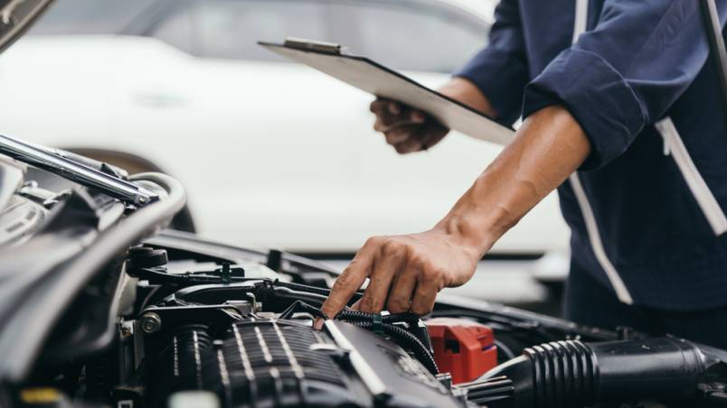 Das Bild zeigt eine Person, die den Motorraum eines Autos überprüft. Sie hält ein Klemmbrett in der einen Hand und untersucht mit der anderen Hand Bauteile im Motor.