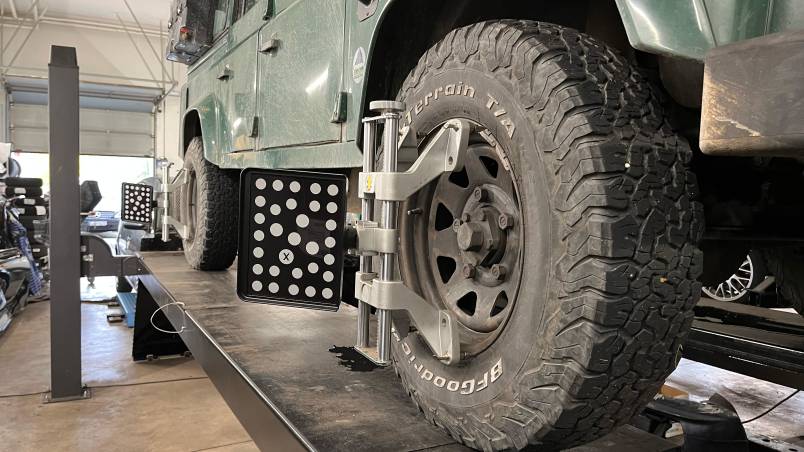 Das Bild zeigt ein grünes Geländefahrzeug auf einer Hebebühne in einer Werkstatt. An den Rädern sind Messgeräte zur Achsvermessung angebracht.